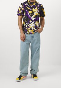 Mångfärgad graphic t-shirt med svart bas, mönster i lila och gult; bärs med ljusblå oversize jeans och färgglada sneakers.