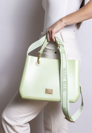 Vrouw met witte broek en top houdt een bleekgroene Marc Ellis handtas met brede verstelbare schouderriem vast.