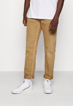 Homme portant un pantalon marron clair retroussé, des chaussettes blanches et des baskets blanches Adidas, debout devant un fond uni clair.