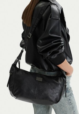 Person trägt eine schwarze Lederjacke und Jeans und trägt eine große schwarze Leder-Schultertasche mit einem markenlogo-bestickten Riemen und Reißverschlussdetail.