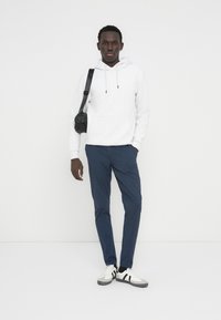 Άνδρας με λευκή φούτερ με κουκούλα, μπλε παντελόνι, λευκά αθλητικά παπούτσια και μαύρη τσάντα χιαστί, στέκεται μπροστά σε απλό φωτεινό φόντο.