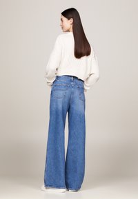 Gekürzter weißer Ripppullover kombiniert mit hoch taillierten, weit geschnittenen blauen Jeans. Die Jeans haben zwei Gesäßtaschen und eine helle Waschung.
