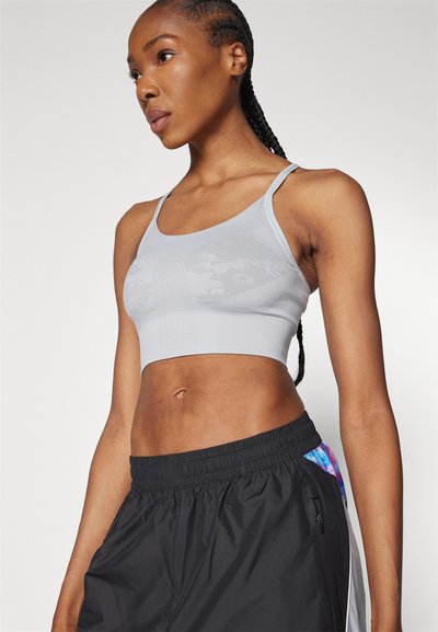 Športovo založená žena s vrkočmi, oblečená v svetlošedej športovej podprsenke a čiernych športových nohaviciach s farebnými bočnými panelmi, pózujúca na jednoduchom pozadí.