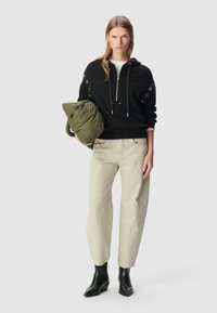 Felpa nera con cappuccio e dettagli ad occhio, giacca leggera imbottita verde oliva, pantaloni beige chiaro e stivali neri alla caviglia.