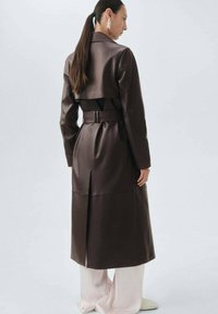 Brązowy sztuczny skórzany płaszcz o kroju trencza z paskiem w talii, rozporkiem z tyłu i wyraźnym kołnierzem. Nosi się go na jasnoróżowych spodniach, prezentując elegancki design.