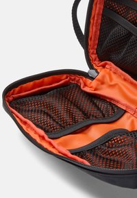 Borsa nera per il trasporto con interno arancione. Presenta tasche in rete, chiusura zip e una finitura in tessuto morbido. Design compatto e funzionale.