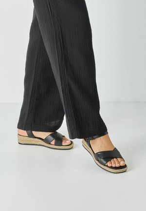 Person trägt schwarze, weit geschnittene Hosen und schwarze Ledersandalen mit gewebten Sohlen auf einem weißen Boden.