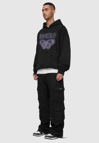 Hanorac negru cu un grafic de fluture violet, asortat cu pantaloni cargo negri ce prezintă multiple buzunare. Ținuta include adidași alb-negru.