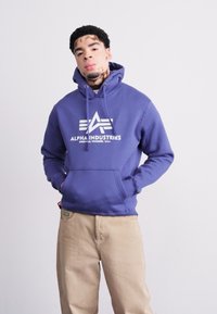 Violetinis džemperis su priekiu kišene ir raišteliais, su balta "Alpha Industries" logo. Dėvimi su smėlio spalvos kelnėmis. Sklandus paviršius.