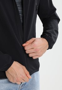 Veste légère noire avec fermeture éclair avant, poignets élastiques et texture lisse. Des mains sont en train de fermer la veste, avec un t-shirt gris en dessous.