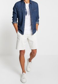 Jeanshemd mit Knöpfen, weißes Grafik-T-Shirt, weiße Shorts und weiße Sneakers. Das Hemd hat zwei vordere Taschen und hochgerollte Ärmel.