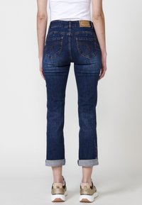 Temno modri kavbojki z zmerno visokim pasom, zavihki na robovih in dekorativnim šivanjem na zadnjih žepih. Nosi se z belimi supergami.