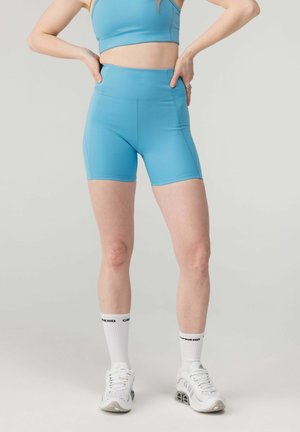 Persoană purtând pantaloni scurți sport albastre și top scurt, șosete albe cu text negru și pantofi sport albi, stând cu mâinile pe șolduri.