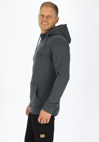 Felpa grigia con tasca frontale, dotata di cappuccio con coulisse, maniche lunghe e una texture morbida. Abbinata a pantaloni neri.