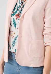 Světle růžový blazer s texturovanou látkou, který má dva knoflíky a kapsu. Pod ním je bílá květinová blůza se zelenými listy.