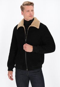 Schwarze Kunstwildlederjacke mit einem beigen Shearling-Kragen, Rei�ßverschluss vorne, gerippten Bündchen und Saum. Modell trägt sie mit einem schwarzen Rollkragenpullover und Jeans.