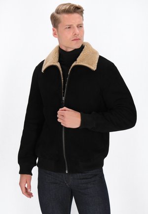 Jachetă din faux suede neagră cu guler din blană de oaie bej, fermoar în față, manșete și tiv cu nervuri. Model purtând-o cu un pulover negru cu guler înalt și blugi.