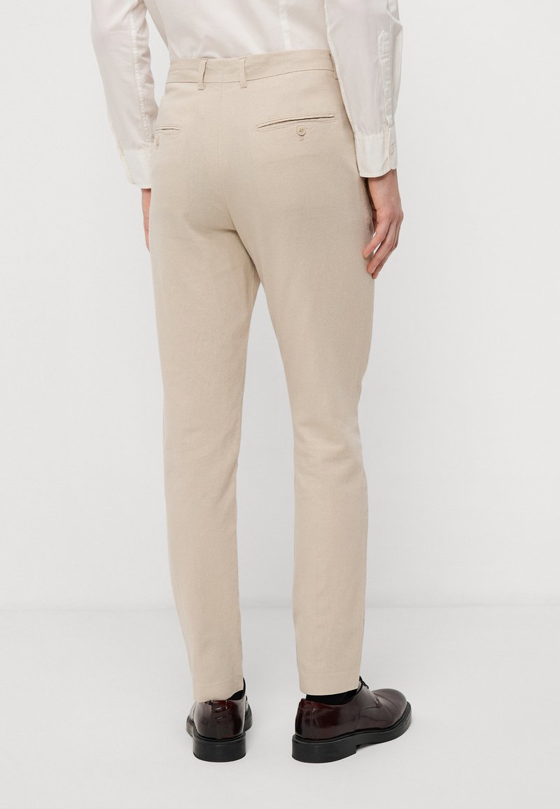 Pantalons beige clair avec une coupe slim, fabriqués en tissu texturé. Dotés de poches arrière avec boutons et portés avec des chaussures marron foncé.