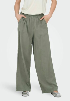 Person iført løstsittende olivengrønne bukser og sølvfargede ballettsko, med hendene tilfeldig plassert i lommene.