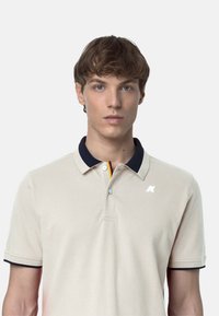 Polo shirt beige con colletto e polsini blu navy. Presenta una chiusura a due bottoni e un logo sul lato sinistro del petto. Materiale in cotone con una texture liscia.