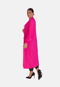 Lång, ljusrosa stickad cardigan med ett strukturerat mönster, öppen framsida och siden kabeldetaljer, parat med svarta högklackade skor och mörka byxor.