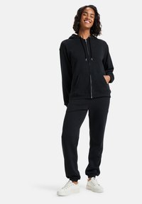 Felpa nera con zip, cappuccio e tasche frontali, abbinata a pantaloni jogger coordinati. Materiale in cotone morbido, con una vestibilità comoda e sneakers bianche.