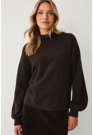 Kvinde med blondt hår iført en mørkebrun langærmet sweater med høj halsudskæring og et mørkt skørt, stående mod en ensfarvet baggrund.