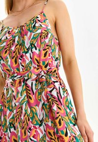 Kukkakuosi mekko, jossa on kirkkaan vaaleanpunaisia, keltaisia ja vihreitä kuvioita valkoisella taustalla. Mekossa on vartalonmyötäinen vyötärö ja nauhakiinnitys. Spagettiolkaimet.