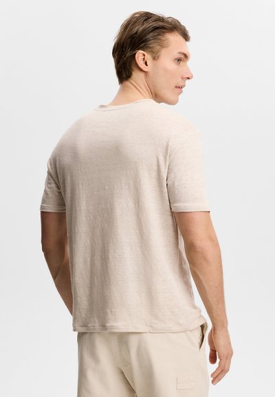 Jeune homme légèrement tourné, portant une chemise beige unie à manches courtes et un pantalon clair sur fond blanc.