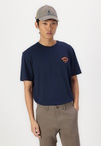 Dunkelblaues Baumwoll-T-Shirt mit kurzen Ärmeln und Rundhalsausschnitt, mit orangefarbenem "Wrangler Jeans 1947" Textlogo. Kombiniert mit hellbraunen Hosen.