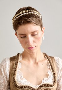Bandeau en perles d'or et d'argent avec des brins entrelacés sur des cheveux courts. Le modèle porte une robe ornée de dentelle avec un décolleté plongeant.