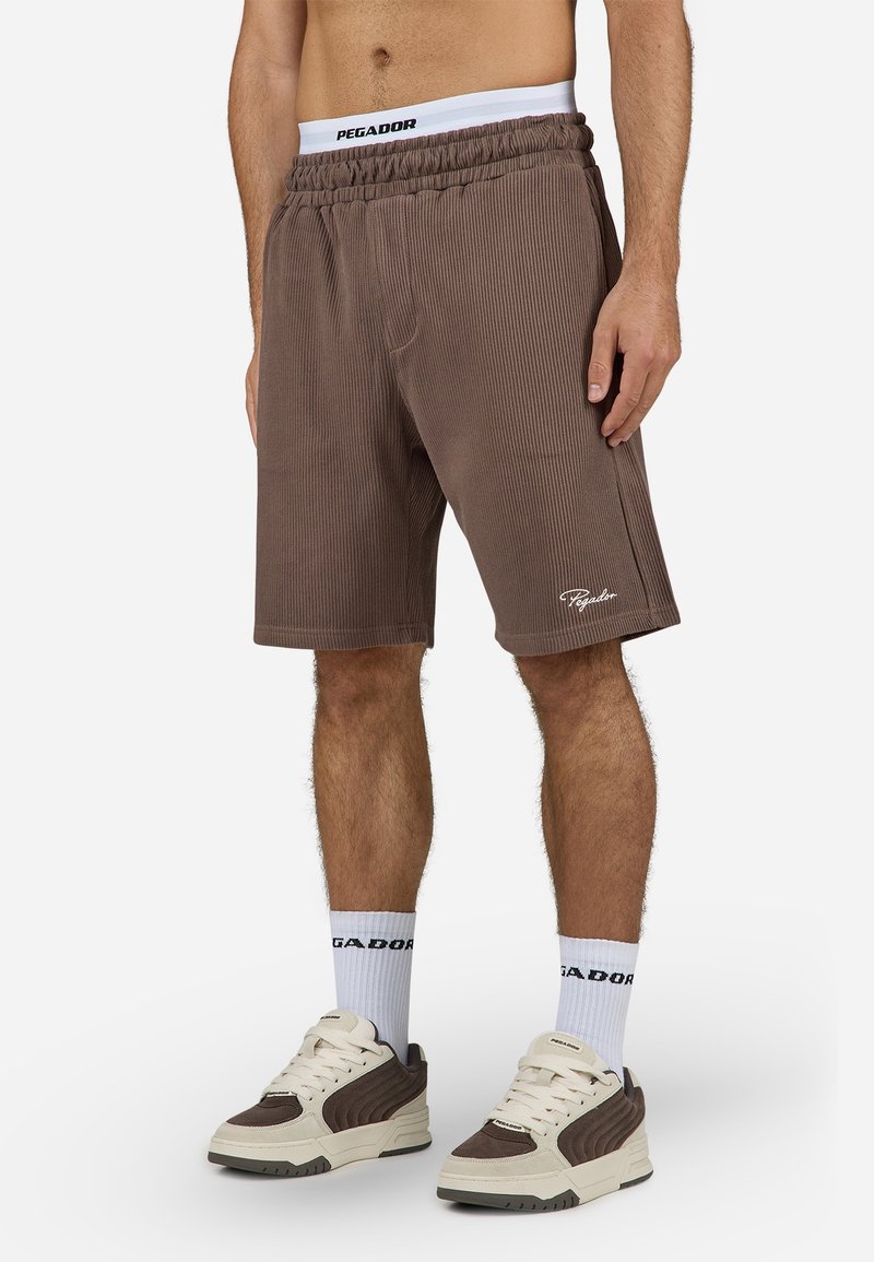 Homme portant un short côtelé marron, des chaussettes blanches de marque et des baskets crème et marron, debout avec une posture détendue sur fond blanc.