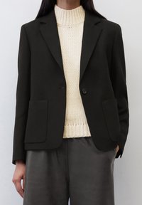 Blazer noir ajusté avec deux poches avant, fermeture à un bouton, texture lisse, assorti à un pull en tricot côtelé crème.