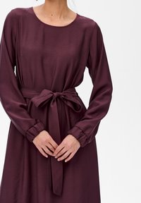 Vrouw in een donkerpaarse jurk met lange mouwen en een stoffen riem in de taille, handen samengevouwen voor zich tegen een witte achtergrond.