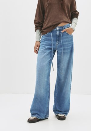 Jeans in denim blu a gamba larga con vita alta, dotati di una zip frontale, tasche e dettagli di cucitura a contrasto. Abbinati a un top marrone.