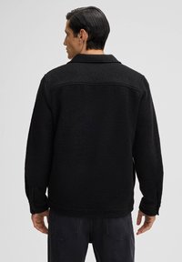 Giacchetto in pile nero con colletto, caratterizzato da un tessuto texture, polsini con bottoni e una vestibilità comoda. La vista posteriore mostra un design semplice e pulito.