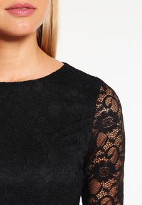 Mustavalkoinen pitsimekko, jossa on pitkät läpinäkyvät pitsihihat. Kaksikerroksinen yläosa kukkakuviolla, pyöreä pääntie ja sileä kangas.