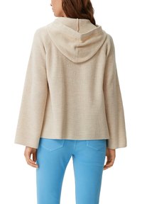 Maglione beige a coste con cappuccio e maniche ampie, caratterizzato da una cucitura posteriore e un cappuccio che pende. Abbinato a jeans aderenti blu chiaro.