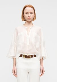 Joven con cabello rojo corto, que lleva una blusa blanca transparente con mangas acampanadas, pantalones blancos y un cinturón marrón con hebilla dorada.