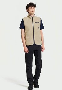 Jovem vestido com um colete de fleece bege sobre uma t-shirt preta, com calças pretas e sapatos pretos, de pé contra um fundo claro e uniforme.