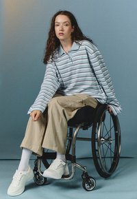 Randig ljusblå långärmad polo med krage, beige vida byxor och vita sneakers. Modell sittande i rullstol.