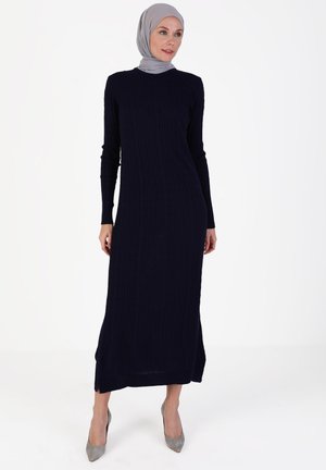 Navy ribbed long dress with long sleeves and a round neckline, featuring side slits and paired with grey high-heeled shoes.