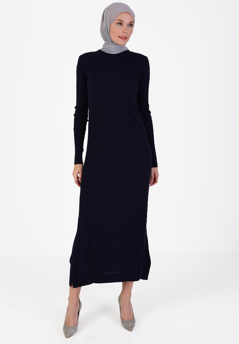 Navy ribbed long dress with long sleeves and a round neckline, featuring side slits and paired with grey high-heeled shoes.