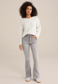 Weißer gestrickter Cardigan mit Rundhalsausschnitt und Knopfverschluss, kombiniert mit hellgrauen Schlagjeans und weißen Sneakers mit braunen Sohlen.