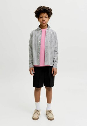 Jeune personne debout portant une chemise à carreaux boutonnée sur un t-shirt rose, un short noir, des chaussettes blanches et des chaussures beiges sur un fond uni.