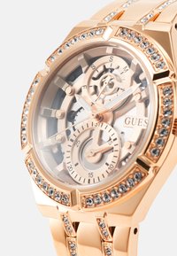 Orologio dorato con quadrante trasparente, ingranaggi intricati e accenti luminosi. Presenta decorazioni in cristallo sulla lunetta e sul cinturino.