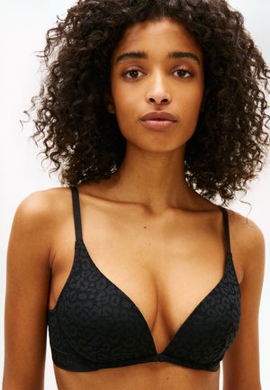 Giovane donna con capelli ricci che indossa un reggiseno nero con stampa leopardata e spalline sottili, rivolta frontalmente su uno sfondo chiaro uniforme.