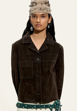 Młoda kobieta ubrana w ciemnobrązową zamszową kurtkę, beżową dzianą czapkę z koralikami i ozdobnymi agrafkami, srebrne wiszące kolczyki oraz turkusowy szydełkowany pasek z dzwoneczkami.