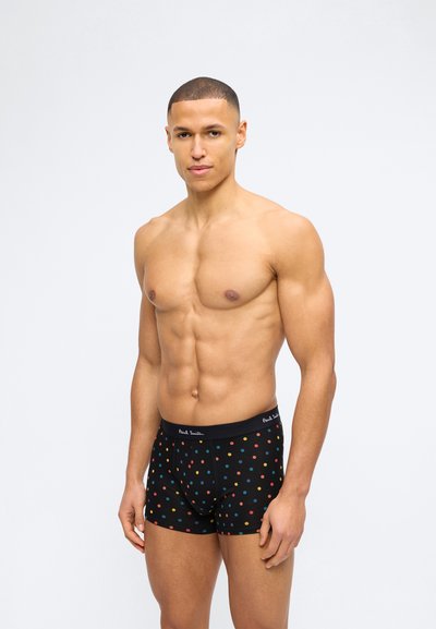 Μυώδης άνδρας που φορά μαύρο βαμβακερό boxer με πολύχρωμες πουά και λάστιχο μέσης με τη λέξη "Paul Smith", στέκεται μπροστά σε λευκό φόντο.