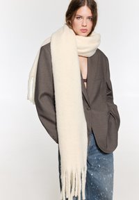 Vit ullsjal med fransade ändar, bärs över en brun oversize blazer. Outfiten inkluderar blå slitna jeans med en slät textur.